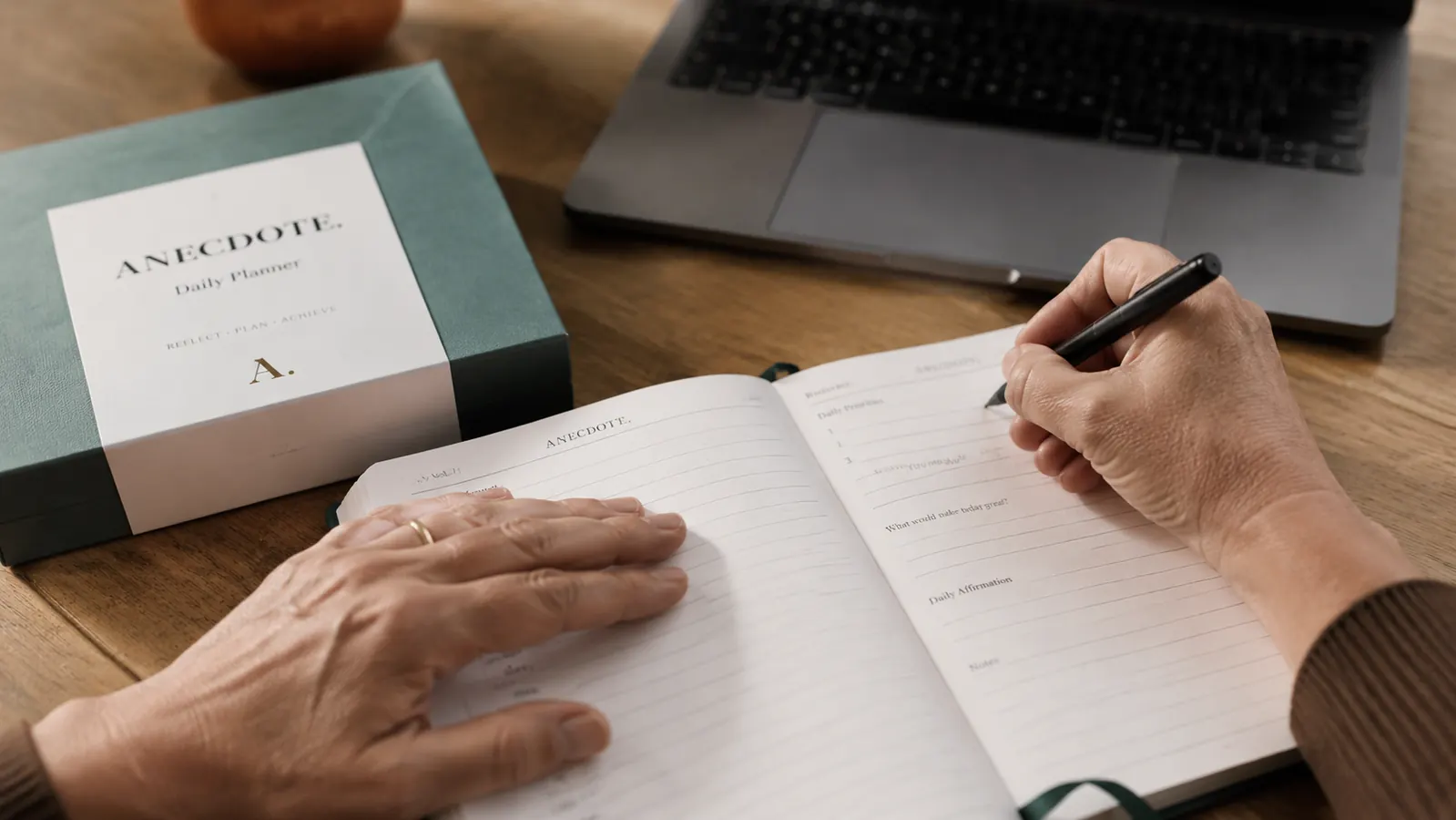 Hand writing morning priorities in the Anecdote Daily Planner on a desk with a laptop in the background
