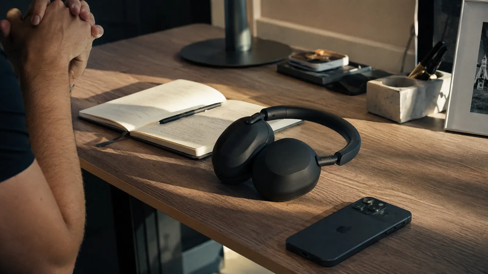 Sony WH-1000XM5 headphones on a standing desk beside a notebook, pen, and smartphone in a modern professional office